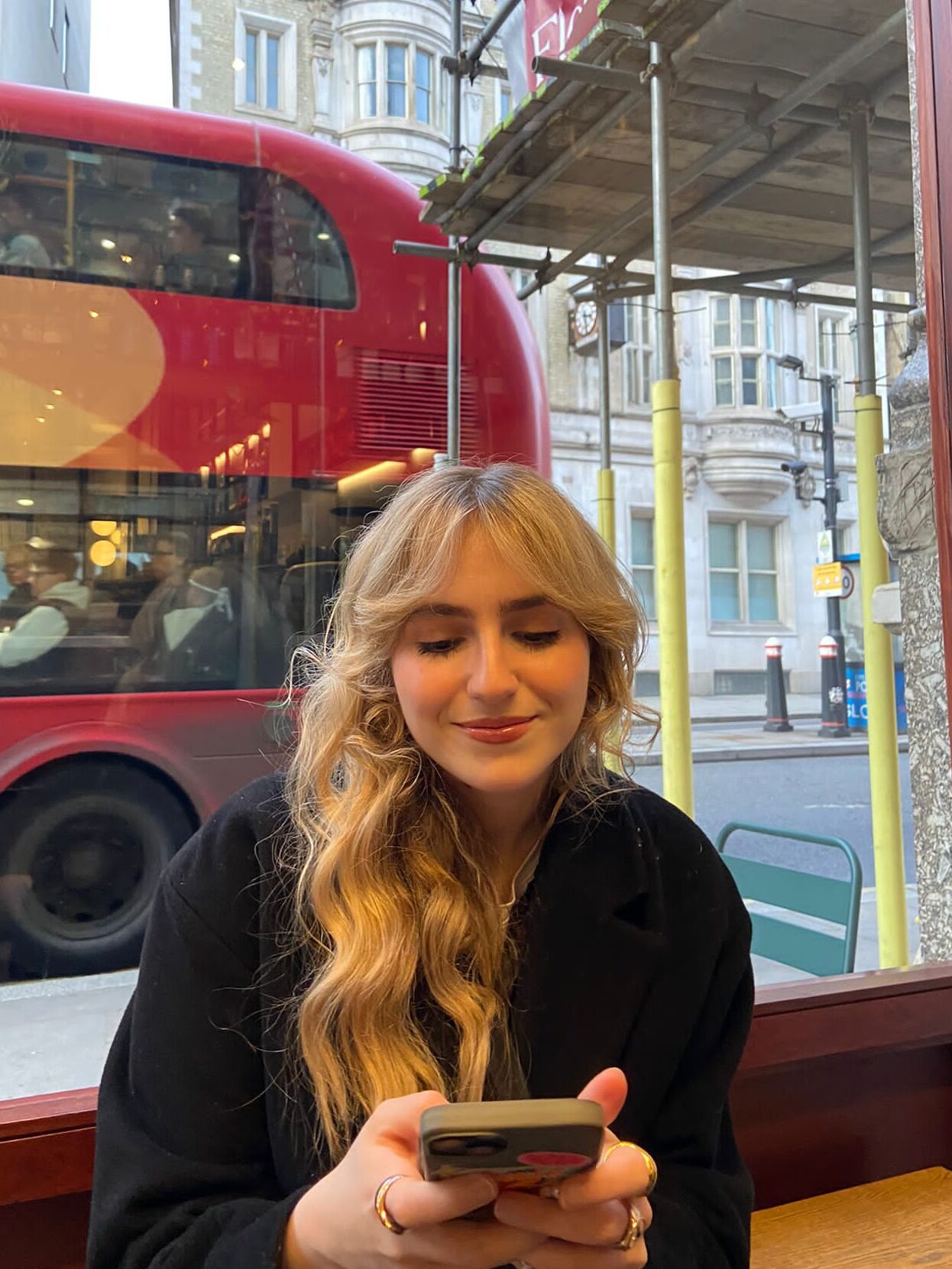 Carmen, a white blonde woman, is sitting in a coffee shop looking down at her phone and smiling.