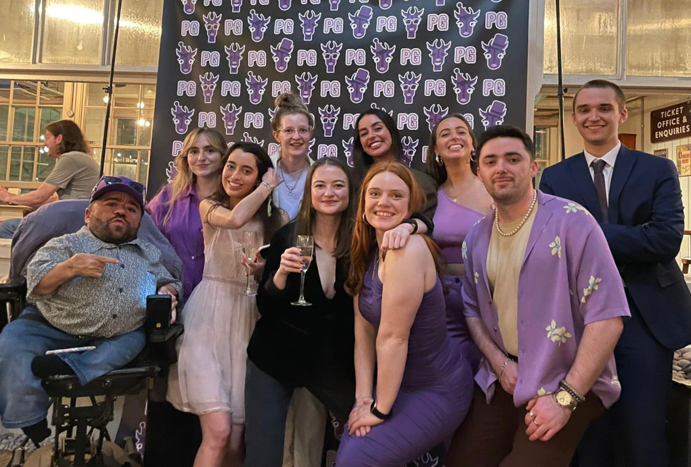 The whole purple goat team except for Martyn Sibley stand in front of a purple goat banner. They are all smiling as they celebrate Purple Goat's second birthday.