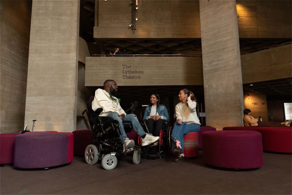 Three friends are in the waiting area together in a theatre. The friends come from diverse backgrounds and the image demonstrates intersectionality. American Express