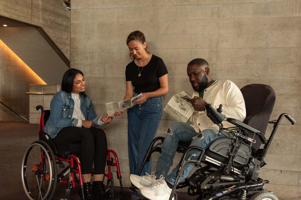 Three friends hold pamphlets as they decide which show to watch at the theatre. The friends come from diverse backgrounds and the image demonstrates intersectionality. American Express
