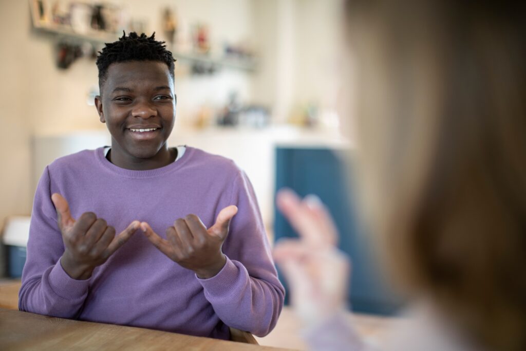 Young man smiling and using sign language in a casual indoor setting, engaging in conversation with another person out of focus in the foreground using a form of Sign Language.