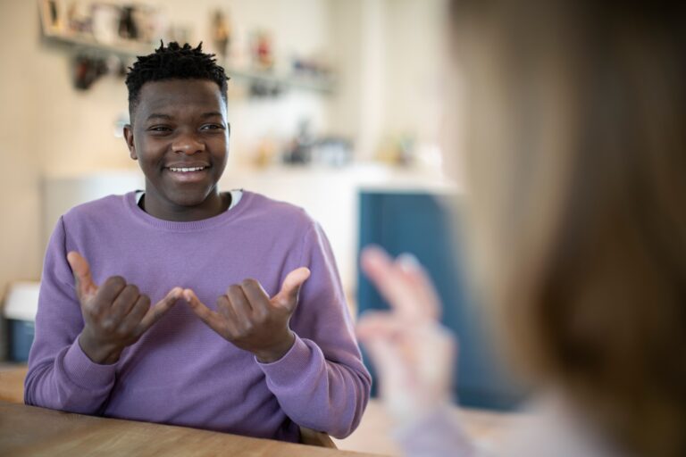 Young man smiling and using sign language in a casual indoor setting, engaging in conversation with another person out of focus in the foreground using a form of Sign Language.