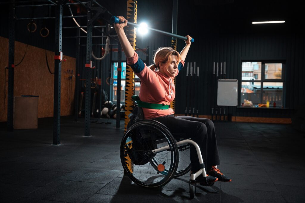A white woman is sitting in her wheelchair in a gym, she's lifting a barbell above her head.