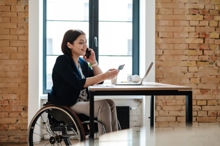 A female wheelchair user is sat in front of her laptop speaking to someone on her phone, while holding a credit card and smiling.
