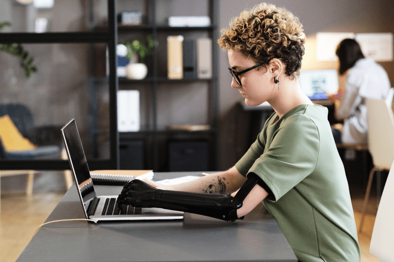 A white person with short blonde hair and glasses, is typing on their laptop. They have a prosthetic arm.