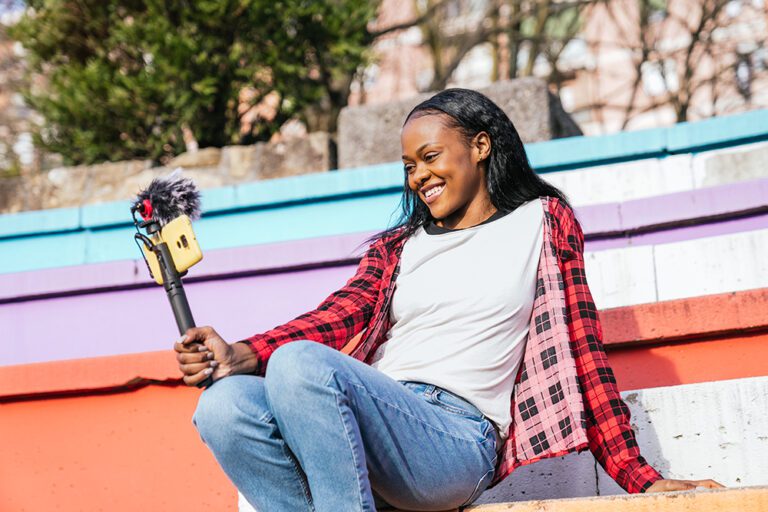 A woman is sat outside on some colourful steps, holding a selfie stick and looking at her phone while she films herself.