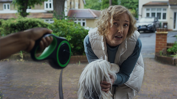Woman stands on a driveway petting a dog. Someone off screen is holding up the dog's lead, and the woman is leaning in to listen to them, unable to hear clearly what they are saying.