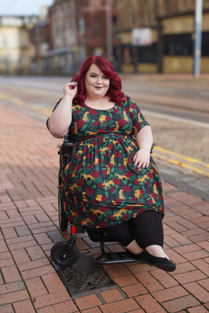 Georgina, a white disabled woman with red hair, is sitting on her wheelchair wearing a colourful dress.
