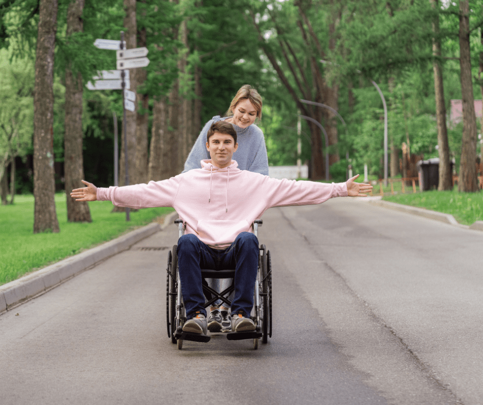 A picture of a young man in a wheelchair with his arms spread out wide, a young woman behind him is smiling. They're in a road surrounded by trees.