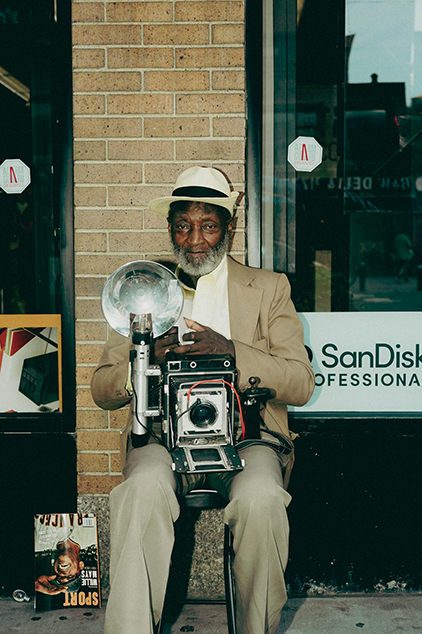 An elderly man sitting outside a shopfront with an old bulb camera.
