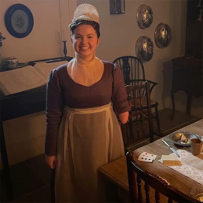 Actress Gracie McGonigal smiling in behind-the-scenes photo of her playing Hazel in Bridgerton. She wears a traditional maid outfit from the early 20th century.