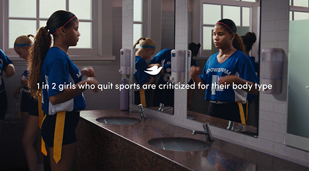 Young girl nervously looks at herself in the mirror in a girl's changing room before going out to play sport. Text on the screen reads: "1 in 2 girls who quit sports are criticized for their body type."