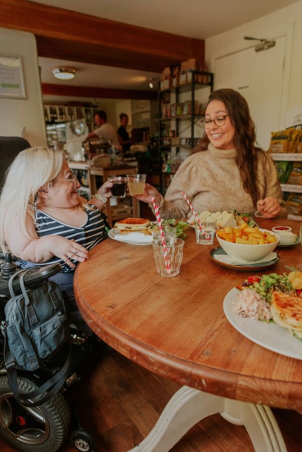 Two women - one blonde with glasses and in a wheelchair, the other brunette with glasses - in a cafe cheersing their drinks. 