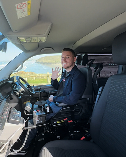 GM-Coachwork campaign image: A powered wheelchair user is positioned in front of the steering wheel in his Wheelchair Accessible Vehicle. He is smiling and waving to the camera.
