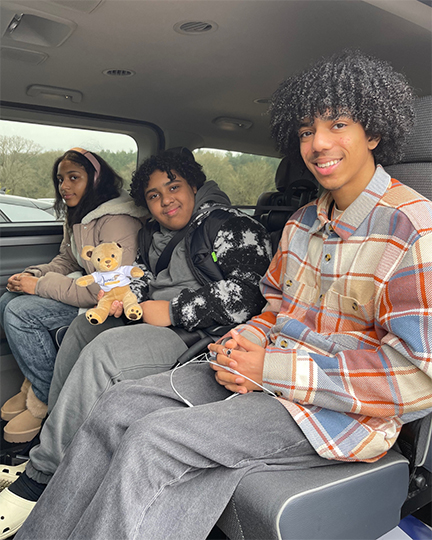 GM-Coachwork campaign image: Three teenagers sit together in the back of a Wheelchair Accessible Vehicle, looking to the camera. The middle child, who is a powered wheelchair user, is holding up a small bear who is used as a mascot by GM Coachwork.