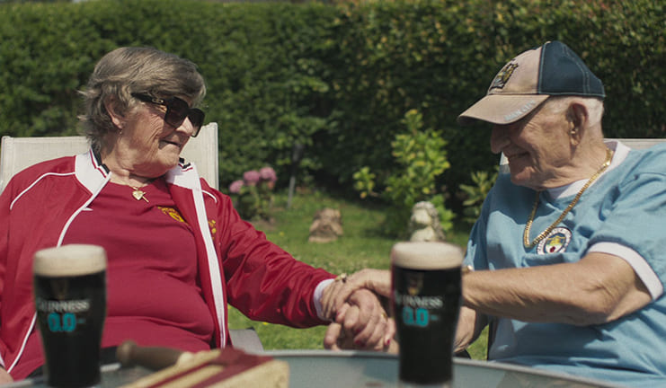 Elderly couple sit in their garden holding hands. Both are wearing football shirts for their favourite teams and they both have a Guinness in front of them.