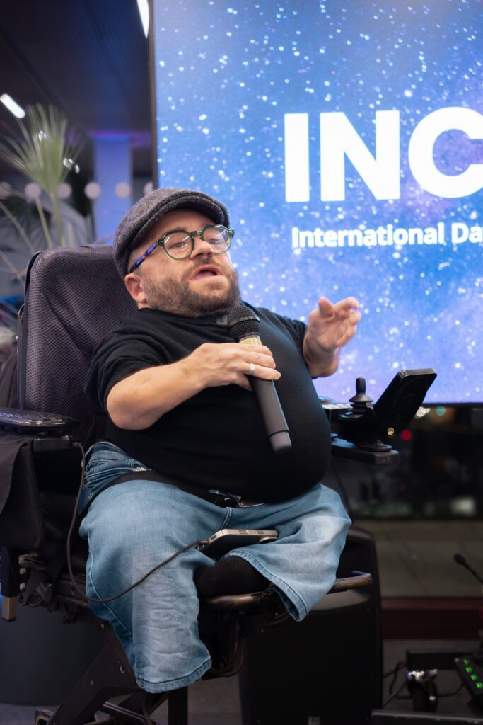An image of Dom, a small white man sitting on his powered wheelchair, talking to an audience and holding a microphone.