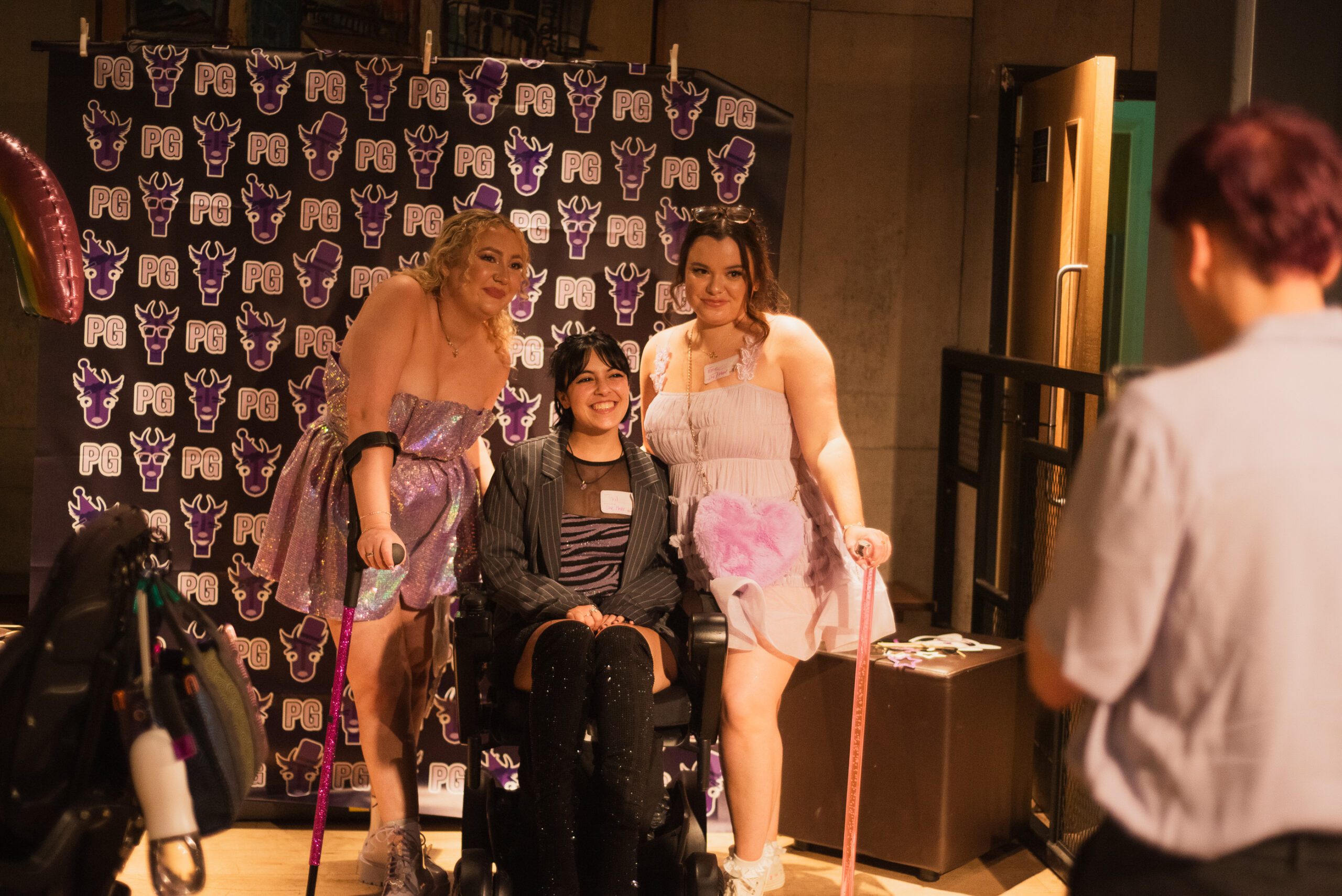 Three disabled people are taking a picture together in front of a backdrop with Purple Goat's logo on it.