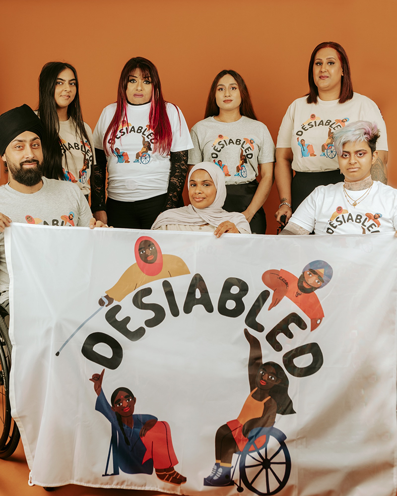 Umaymah poses with other South Asian disabled creators. They are holding up a banner with the word "Desiabled," on it with illustrations of disabled South Asian people surrounding it.
