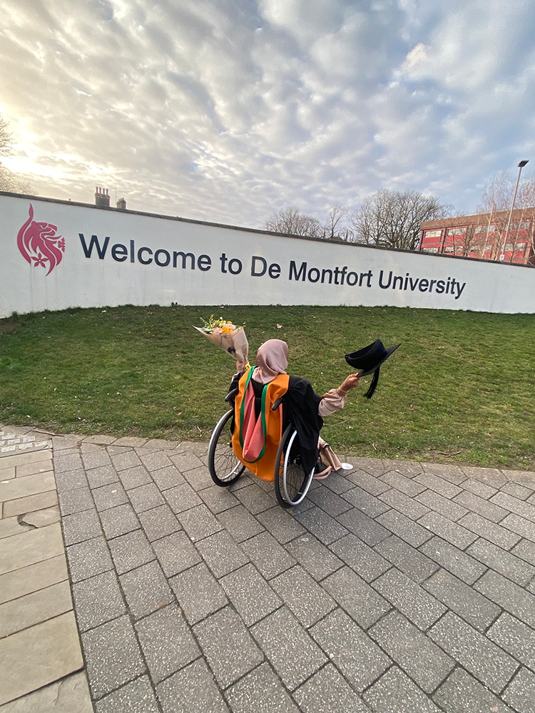 Umaymah celebrates graduating in front of the De Montfort University sign while folding a bouquet of flowers.