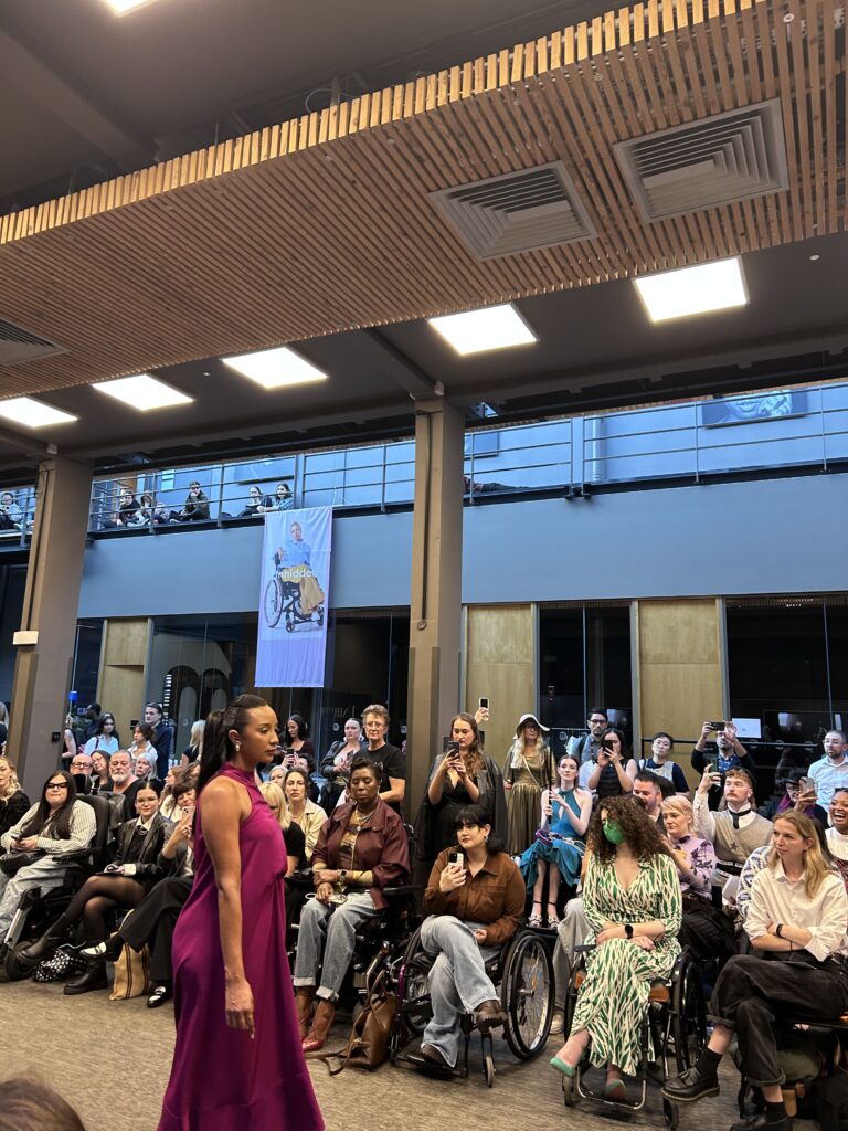 A woman wearing a burgundy coloured dress walking down an inclusive fashion catwalk with a crowd of people with diverse disabilities watching and filming.