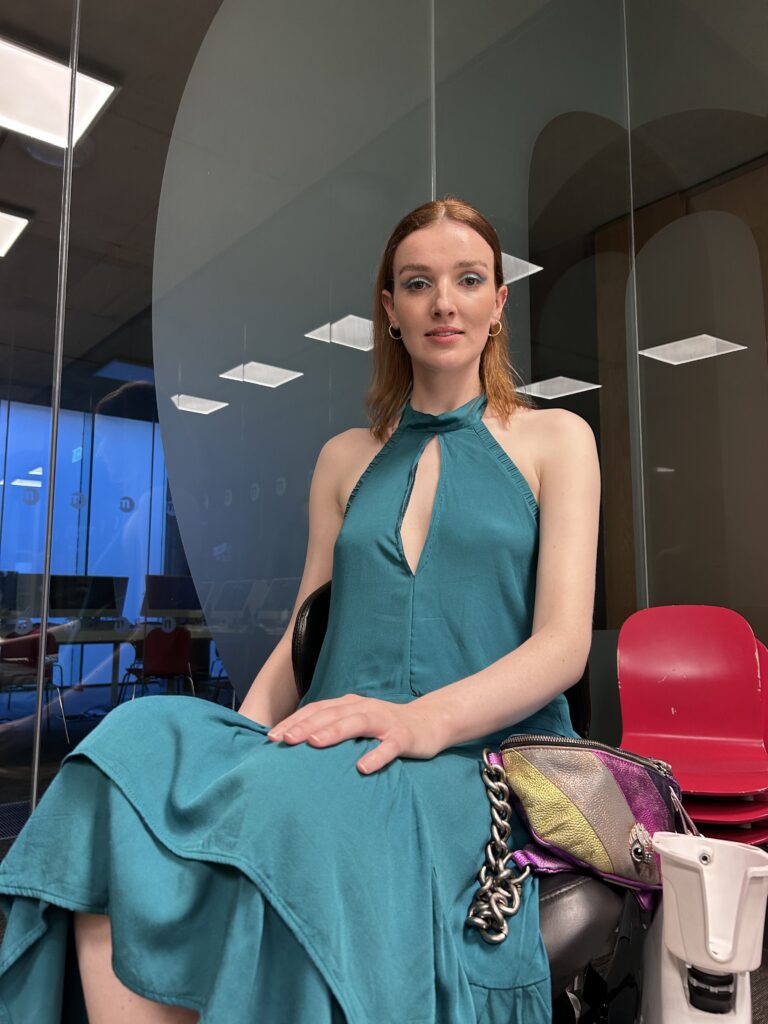 An inclusive fashion model sitting in her wheelchair and looking at the camera, wearing a blue/green dress.