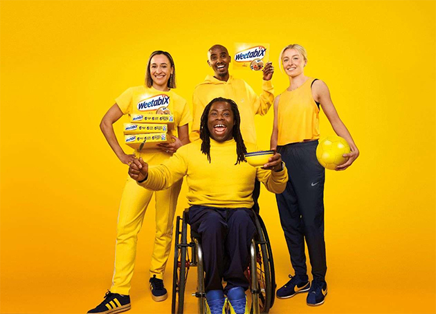 Ade Adepitan and Olympics athletes all hold Weetabix products. They are wearing yellow and dark blue clothes, and are set against a yellow background, to match the Weetabix brand.