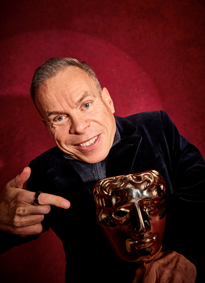 Warwick Davis wearing a formal suit pointing to a BAFTA award that he is holding.