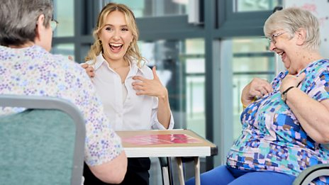 Rose sits at a table with two elderly women. They are all laughing as they practice British Sign Language.