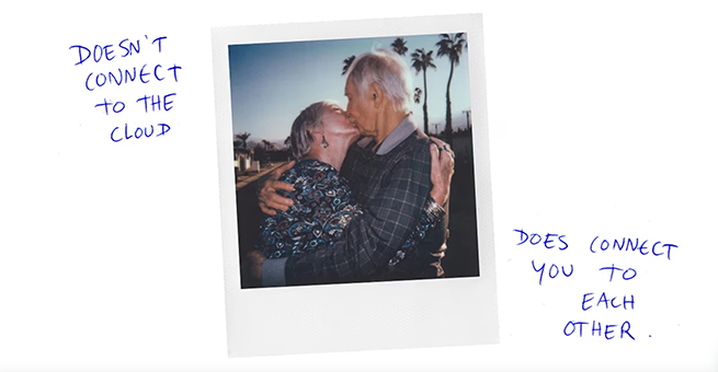 Polaroid photograph of an elderly couple embracing in a coastal, suburban area as the sun sets. Text written in biro around the edge reads: "Doesn't connect to the cloud. Does connect you to each other."