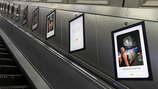 London Underground escalator with posters on the wall showing a photo of a woman at a fireworks event, with text underneath that says: "#ShareTheFirst."