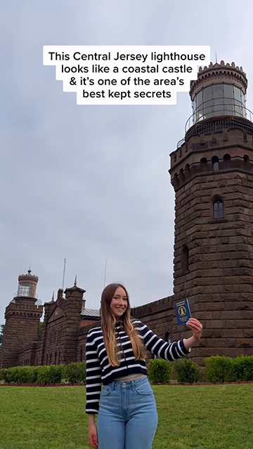 For U.S. Lighthouse Society Campaign, Woman holds up her Lighthouse Passport outside of a lighthouse. Text reads: "This Central Jersey lighthouse looks like a coastal castle & it's one of the area's best kept secrets."