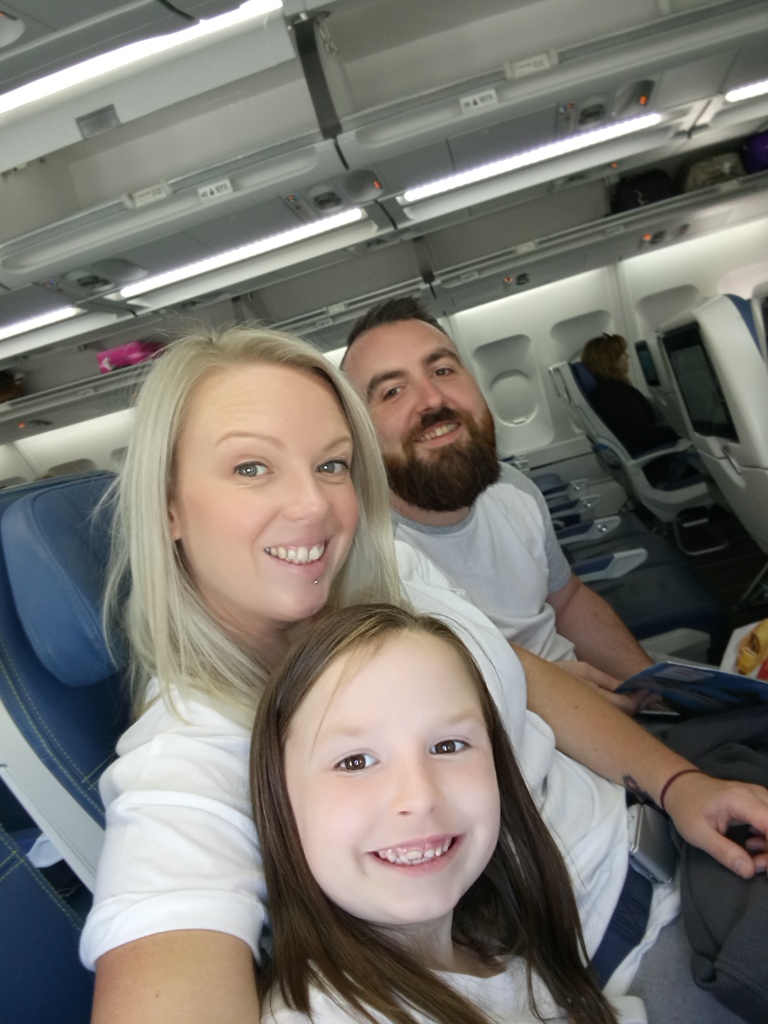Joe, a white man with short brown hair, is sitting on a plane next to her wife and daughter. They're all smiling at the camera.
