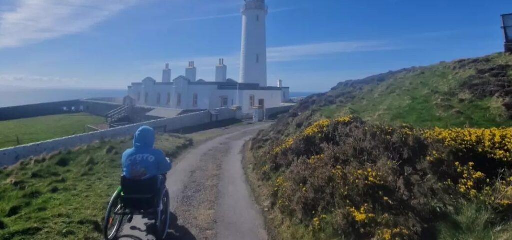 Joe, a male manual wheelchair user, approaches a lighthouse in a hilly, coastal area.