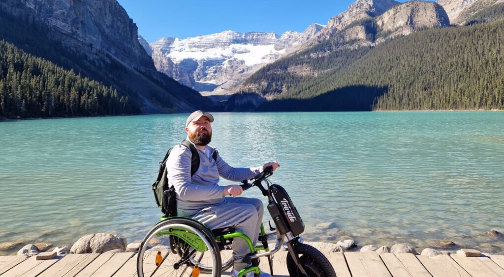 Joe, a white man with dark facial hair, is sitting on his wheelchair. There's a beautiful landscape with a lake and mountains behind him.