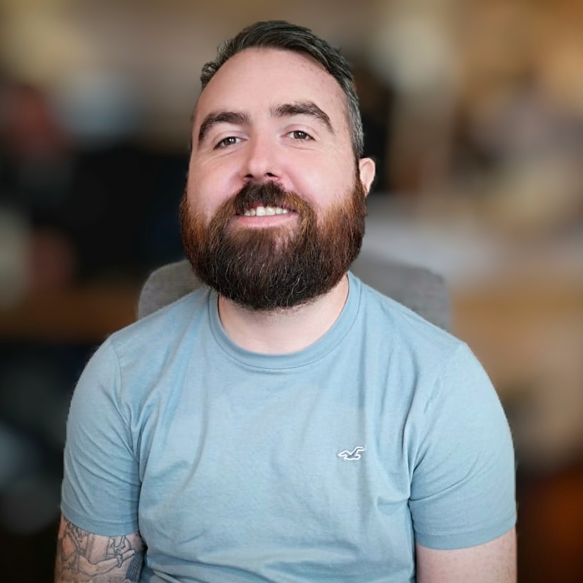 A headshot of Joe, a white man with short brown hair and brown beard. He's smiling at the camera and wearing a blue t-shirt.