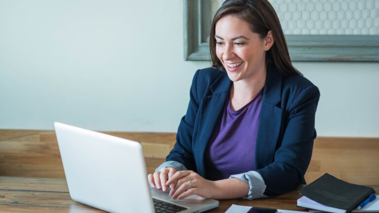 White woman in smart black blazer sat on a laptop smiling.