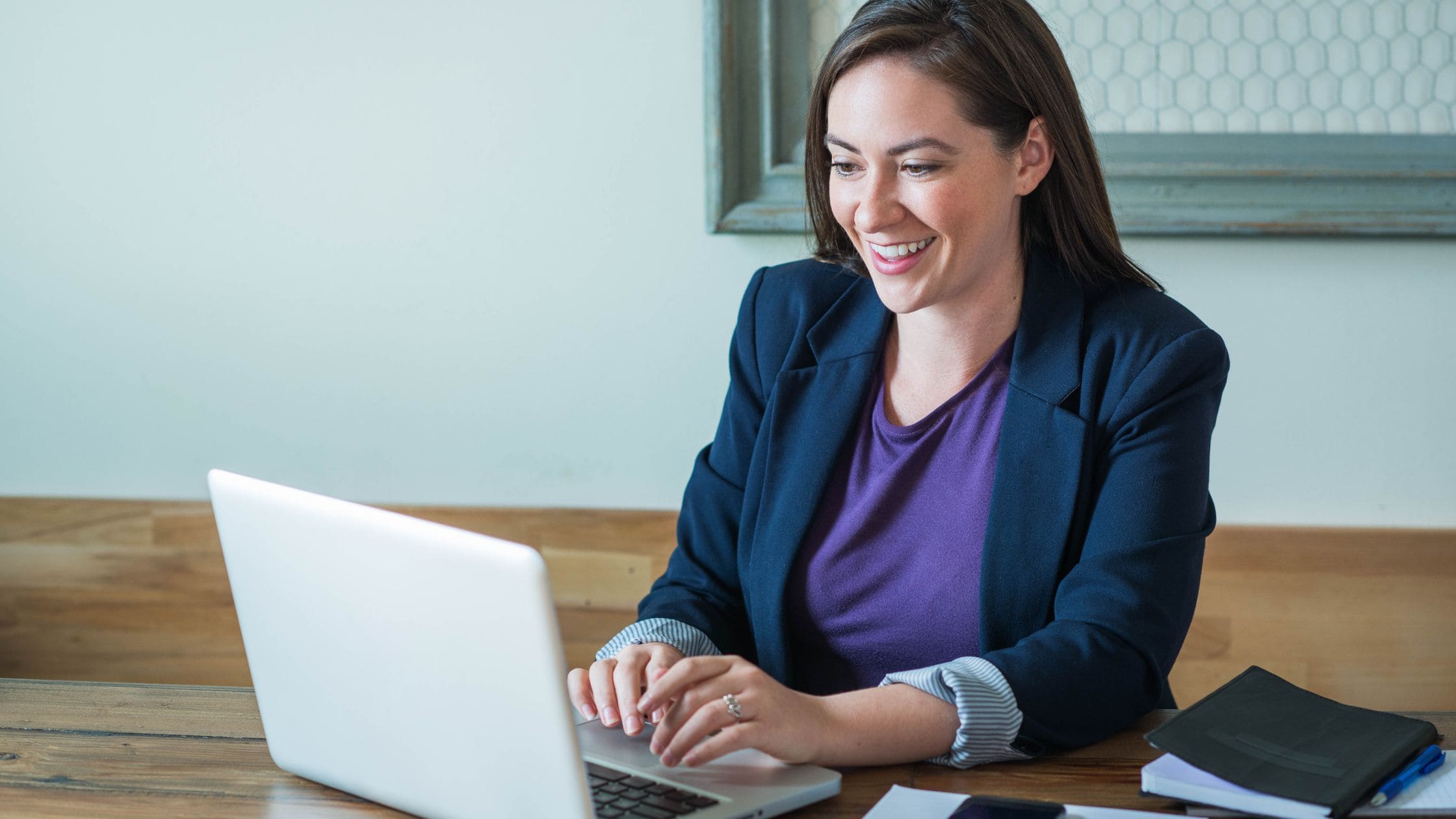 White woman in smart black blazer sat on a laptop smiling.
