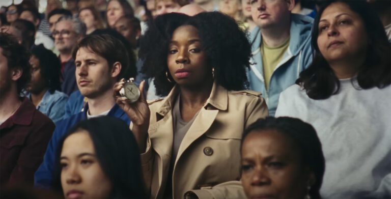 A woman in the audience of a stadium crowd looks to the camera holding a stopwatch with a serious expression on her face.