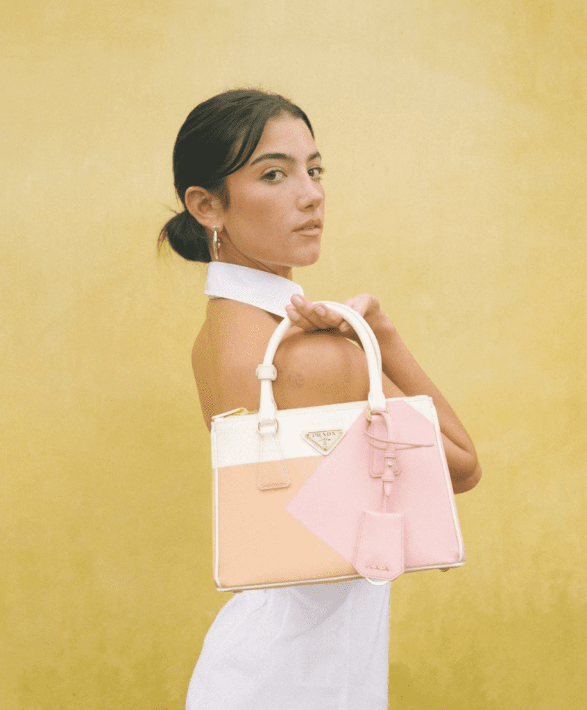 A picture of Charlie, a white young woman with dark hair tied in a low bun. She's wearing a white dress and holding a white and pink bag with the Prada logo on it. The background of the picture is yellow.