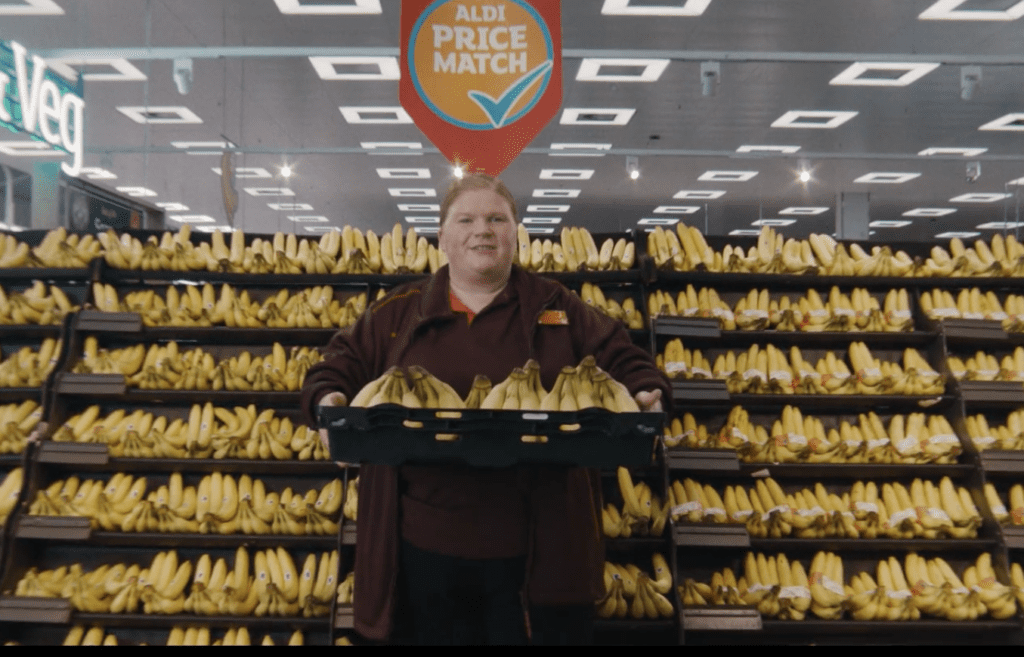 A screenshot from the Sainsbury's ad showing a member of staff in front of a shelf full of bananas, and holding a box full of fruit. Behind them, a sign that says 'Aldi price match'.