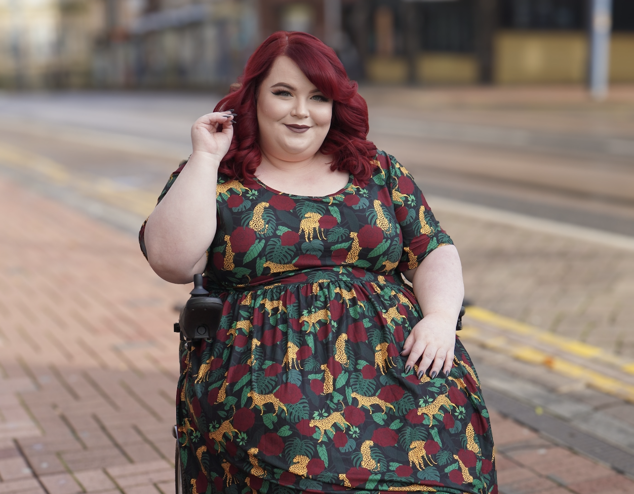 Georgina, a white woman with red hair, is wearing a colourful dress, while sitting in her wheelchair.