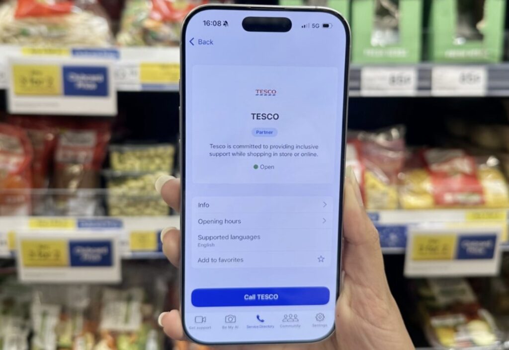 Picture of a phone in front of a food shelf at Tesco, with the 'be my eyes' app opened. Some of the text on the screen says 'Tesco is committed to providing inclusive support while shopping in store or online'.