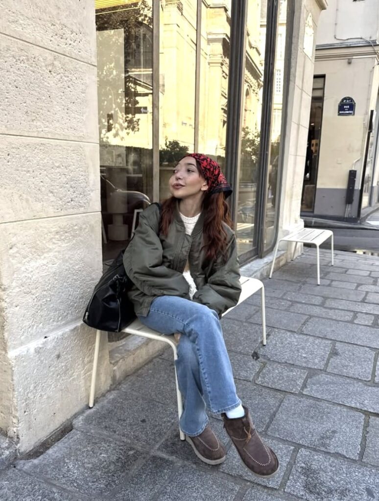 Nikki Lilly, a young woman with a facial difference, is sitting on a bench outside a shop.