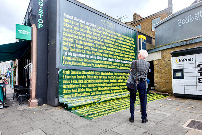 An out-of-home advertisement in a billboard on a street with text that reads "we check check check your chicken," followed by a long list of every chicken product that Waitrose sells. This list is so long that it extends out the bottom of the billboard. A member of the public admires this work up close.