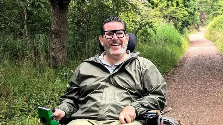 Photograph of Nick Wilson outside in a forested area, looking to the camera and smiling.