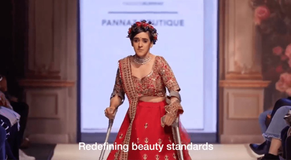 Zubee, a South Asian woman with crutches, is walking down the catwalk of the National Asian Wedding Show, wearing some traditional wedding dress.