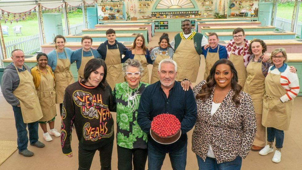 The 12 contestants of the new season of the Great British Bake Off, pose together with the three judges and the host in front of them. One of them is holding a cake. 
