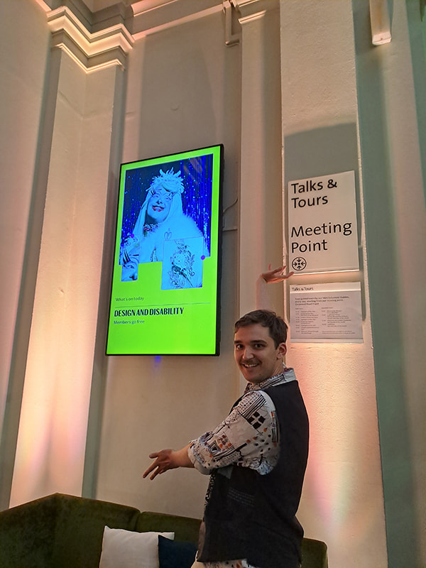 Luke, the article's author, has his arms raised so they frame one of the digital posters for the Design and Disability Exhibition. He looks happily at the camera, proud of the work he helped to make.