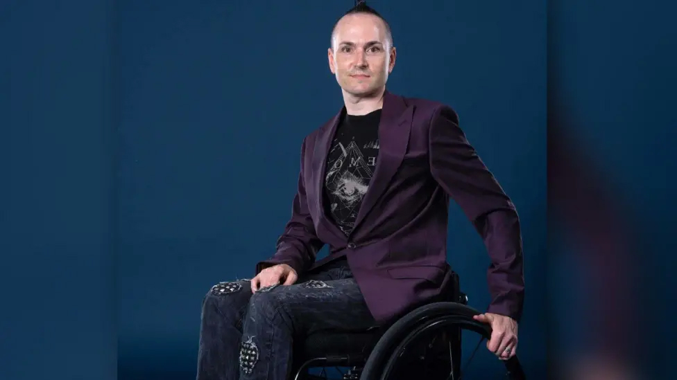 Owen Tooth, a manual wheelchair user, poses in front of a navy blue background for the camera.
